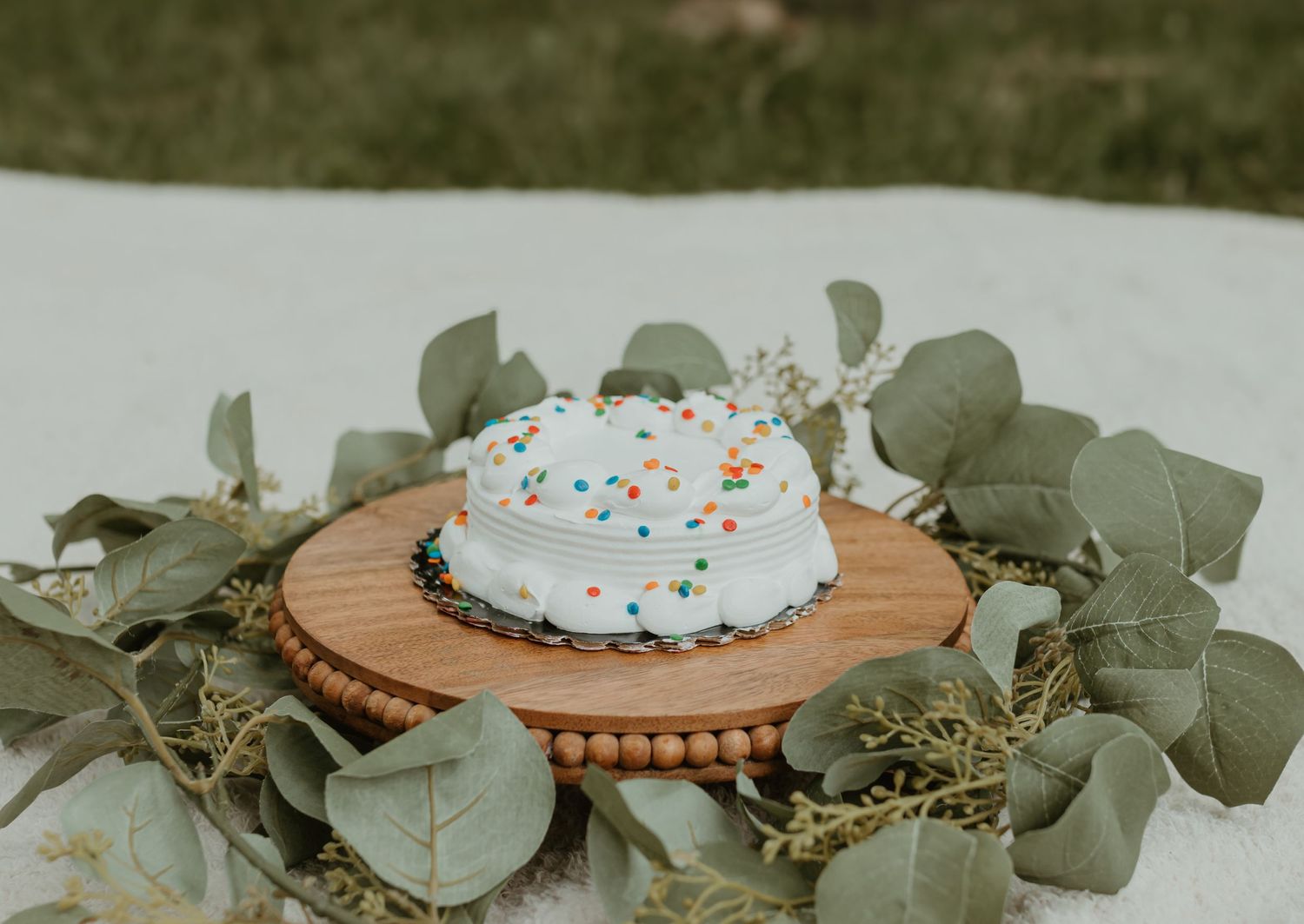 A white frosted cake with sprinkles sits on a wooden cake stand surrounded by eucalyptus leaves.Colorado Springs Photographer