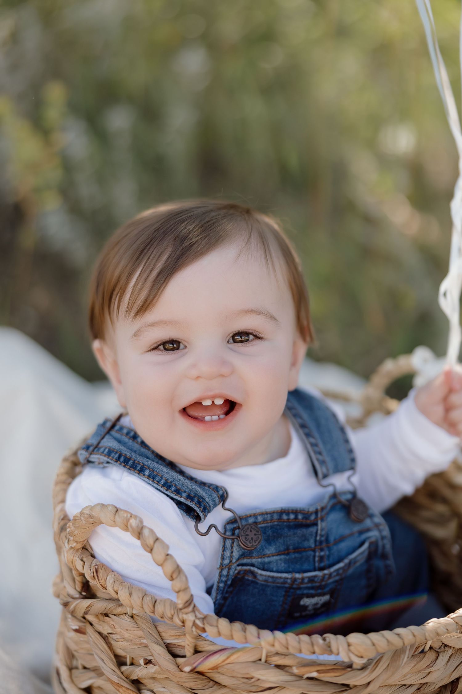 A smiling baby wearing denim overalls and white shirt sits in a woven basket outdoors.