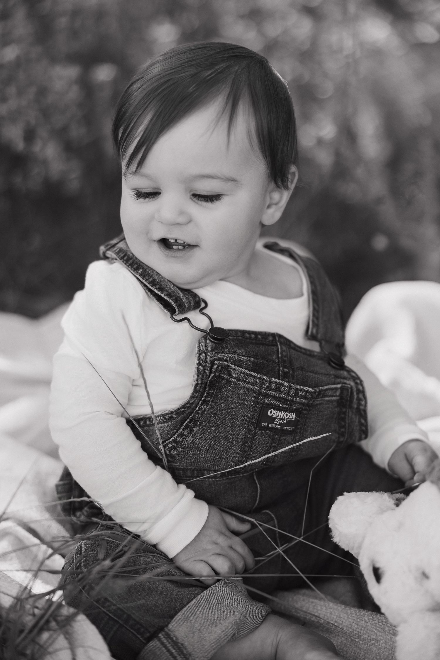 A toddler in denim overalls and white sweater rests peacefully on a blanket in black and white photography.