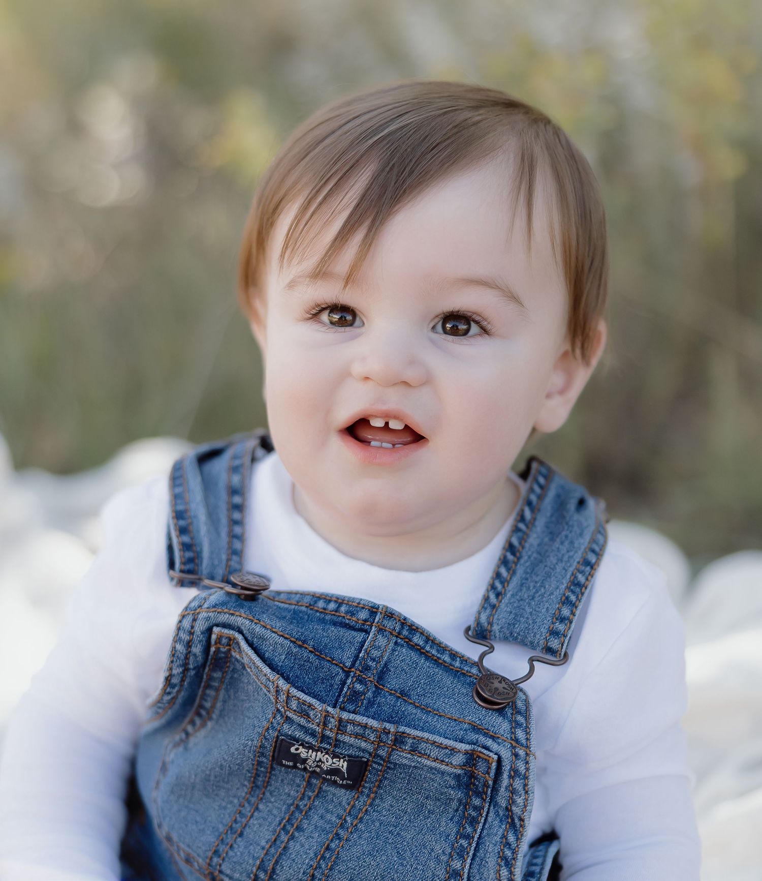 Baby wearing denim overalls and white shirt smiles while sitting outdoors in natural lighting.