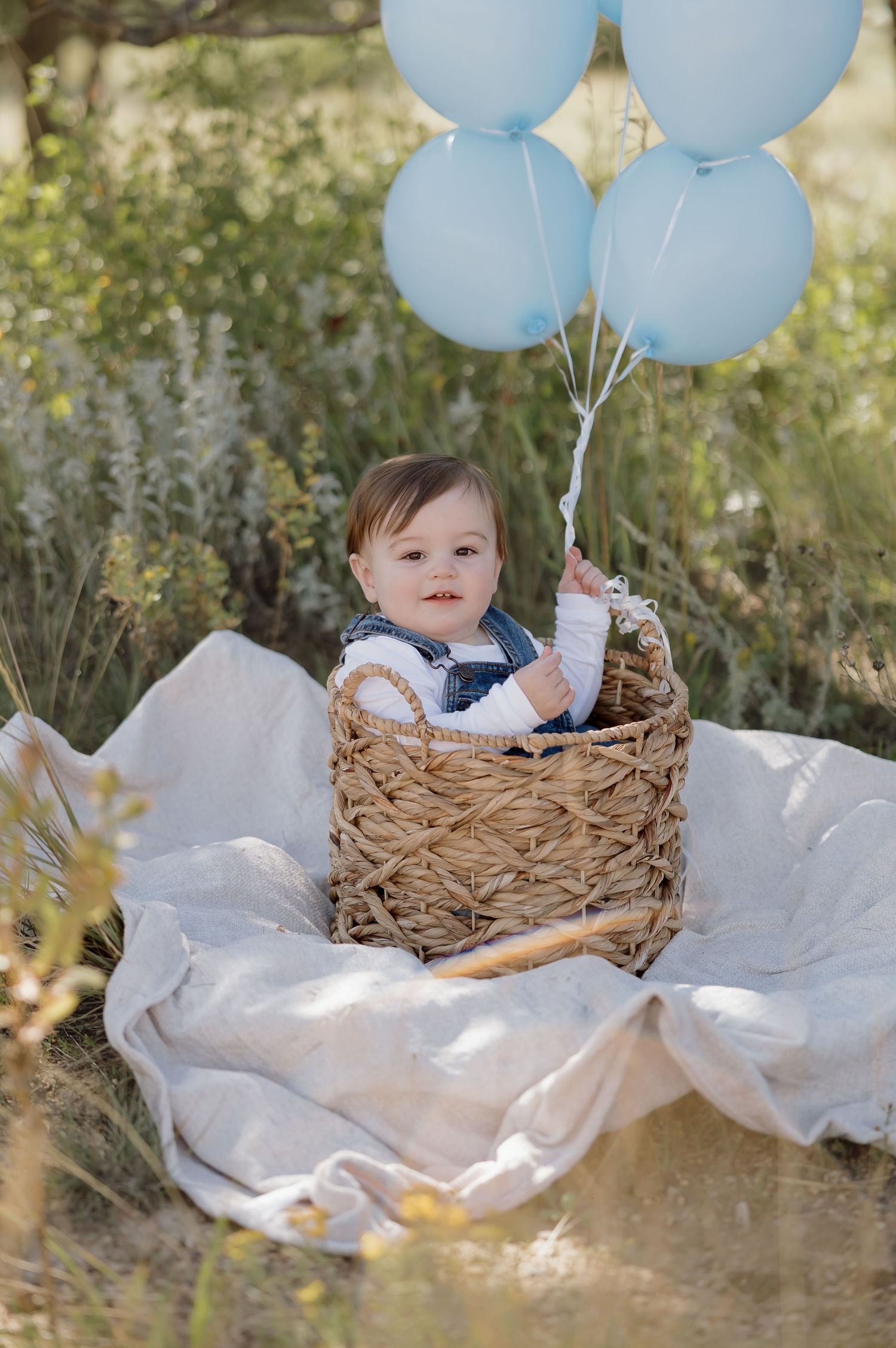 Light blue balloons float above a woven basket set on white fabric in an outdoor garden setting.