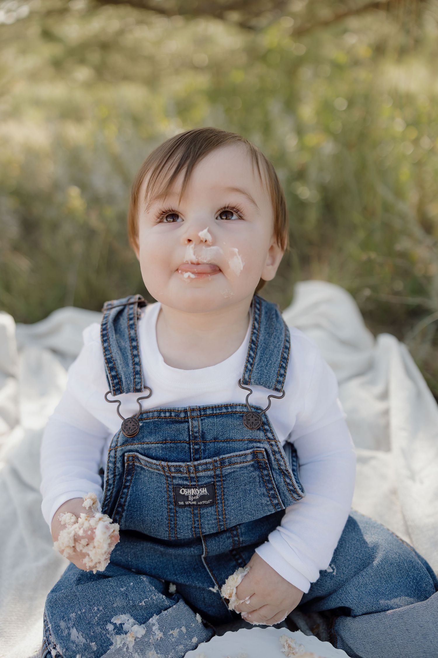 Small child in denim overalls and white shirt sitting outdoors with messy face and hands.
