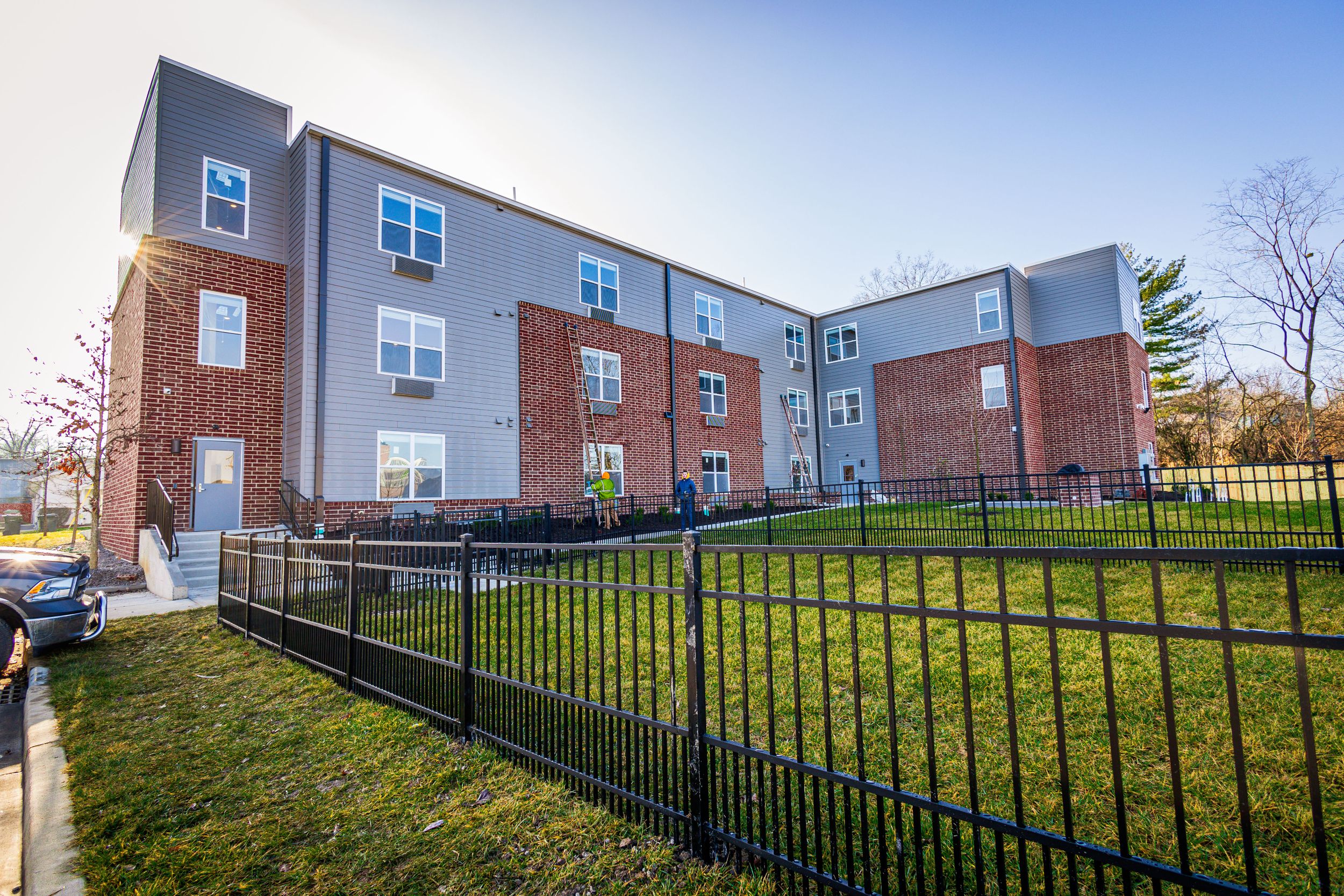 Three-story apartment building with brick and gray exterior featuring a fenced grass courtyard.