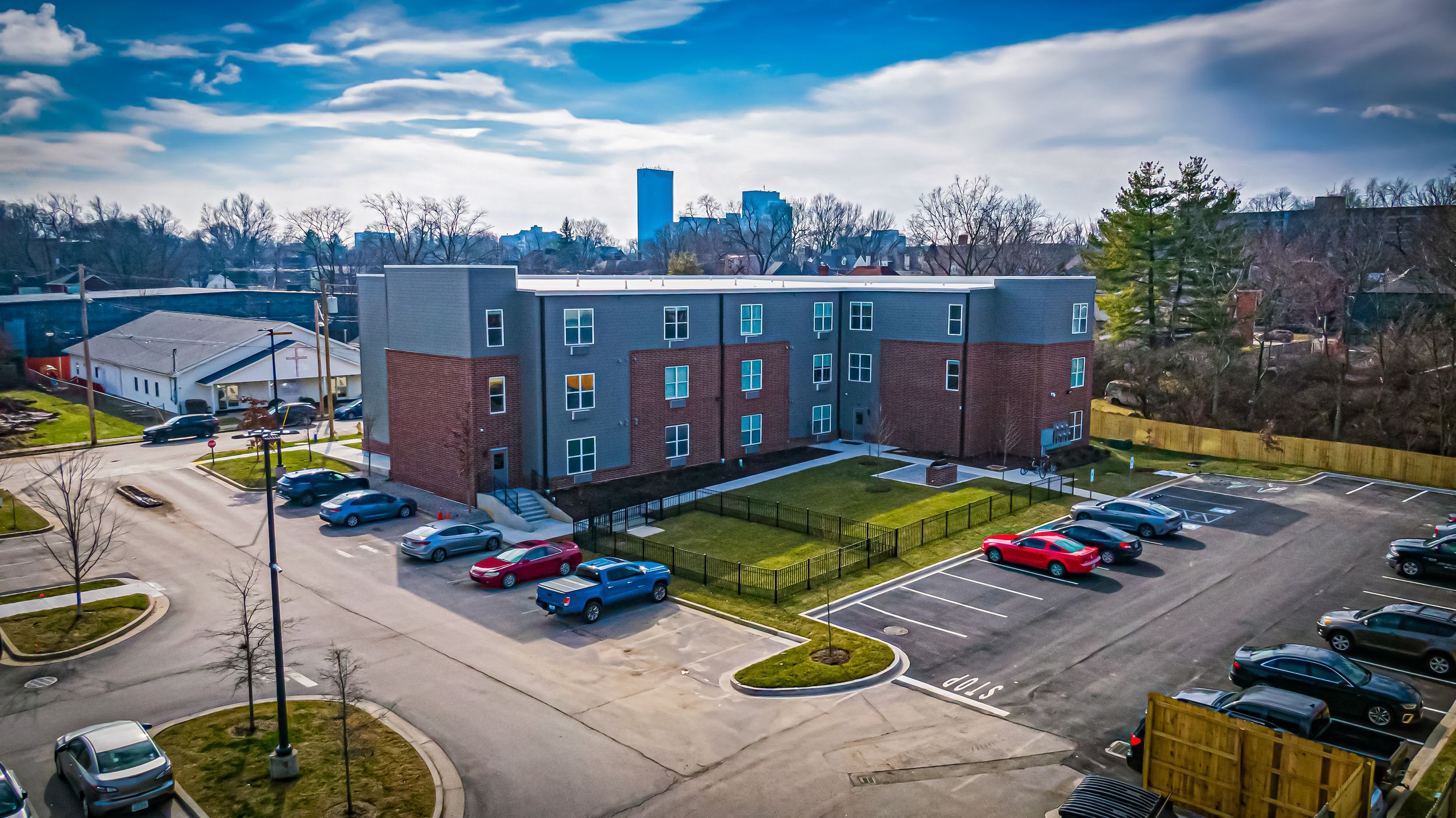 Modern brick apartment complex with blue accents surrounded by parked cars and winter trees.