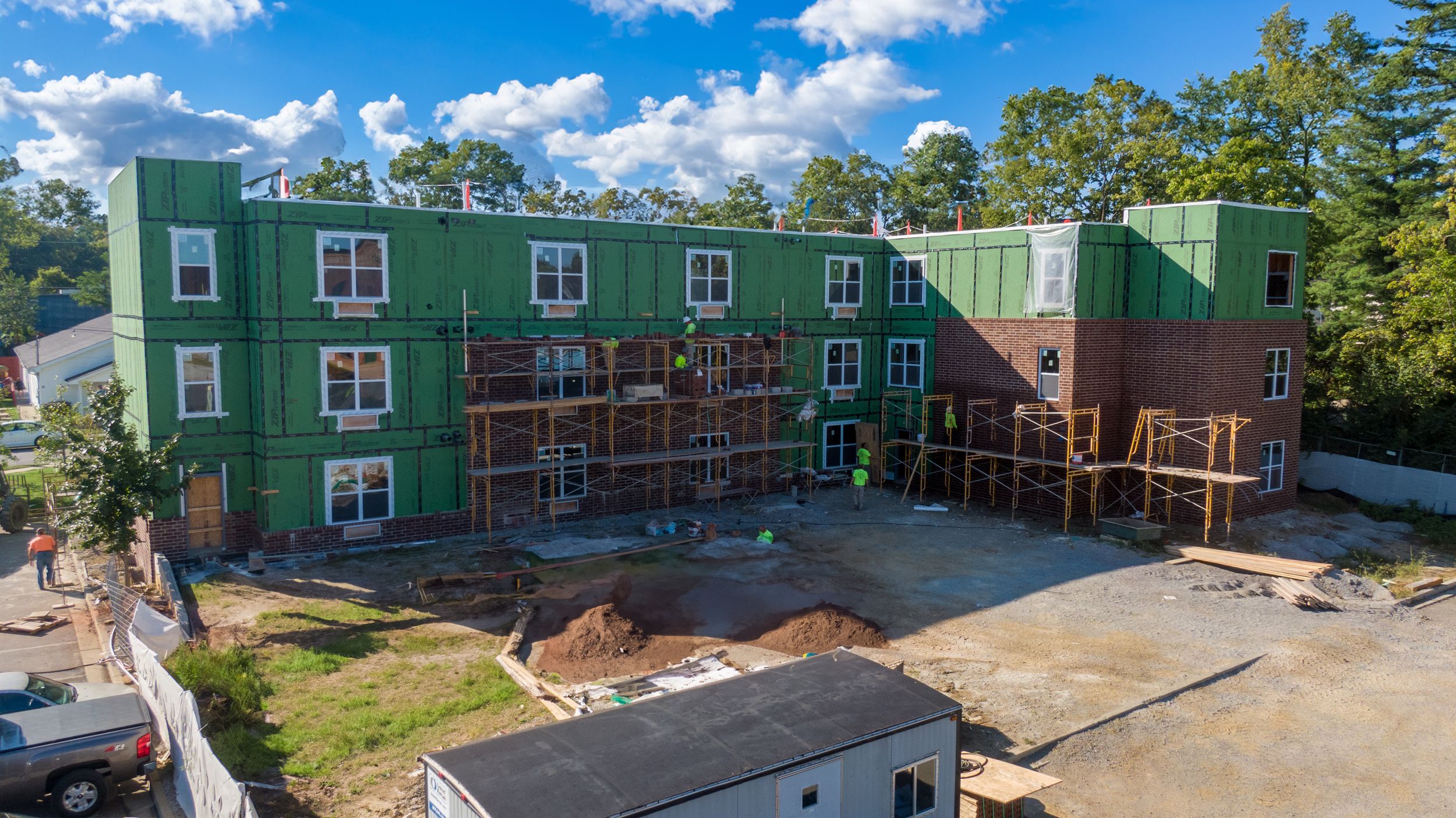 Aerial panorama showing construction progress of green residential apartment buildings surrounded by trees and dirt roads.