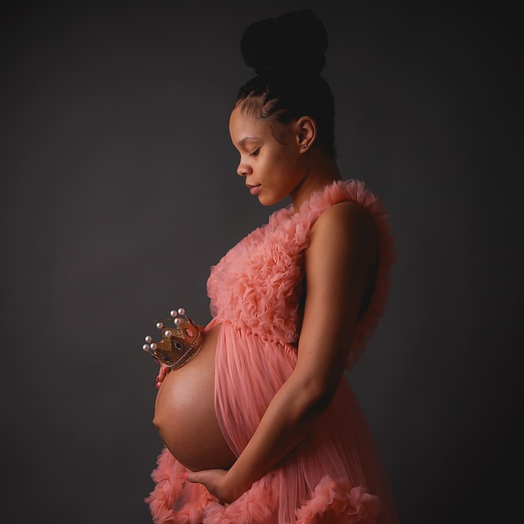 Pregnant woman in a pink ruffled tulle gown gazes down at her belly, holding a small crown in a studio maternity portrait.