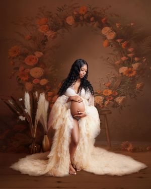 Pregnant woman in a flowing cream ruffled gown sits beneath an orange floral arch with pampas grass in a warm maternity portrait.
