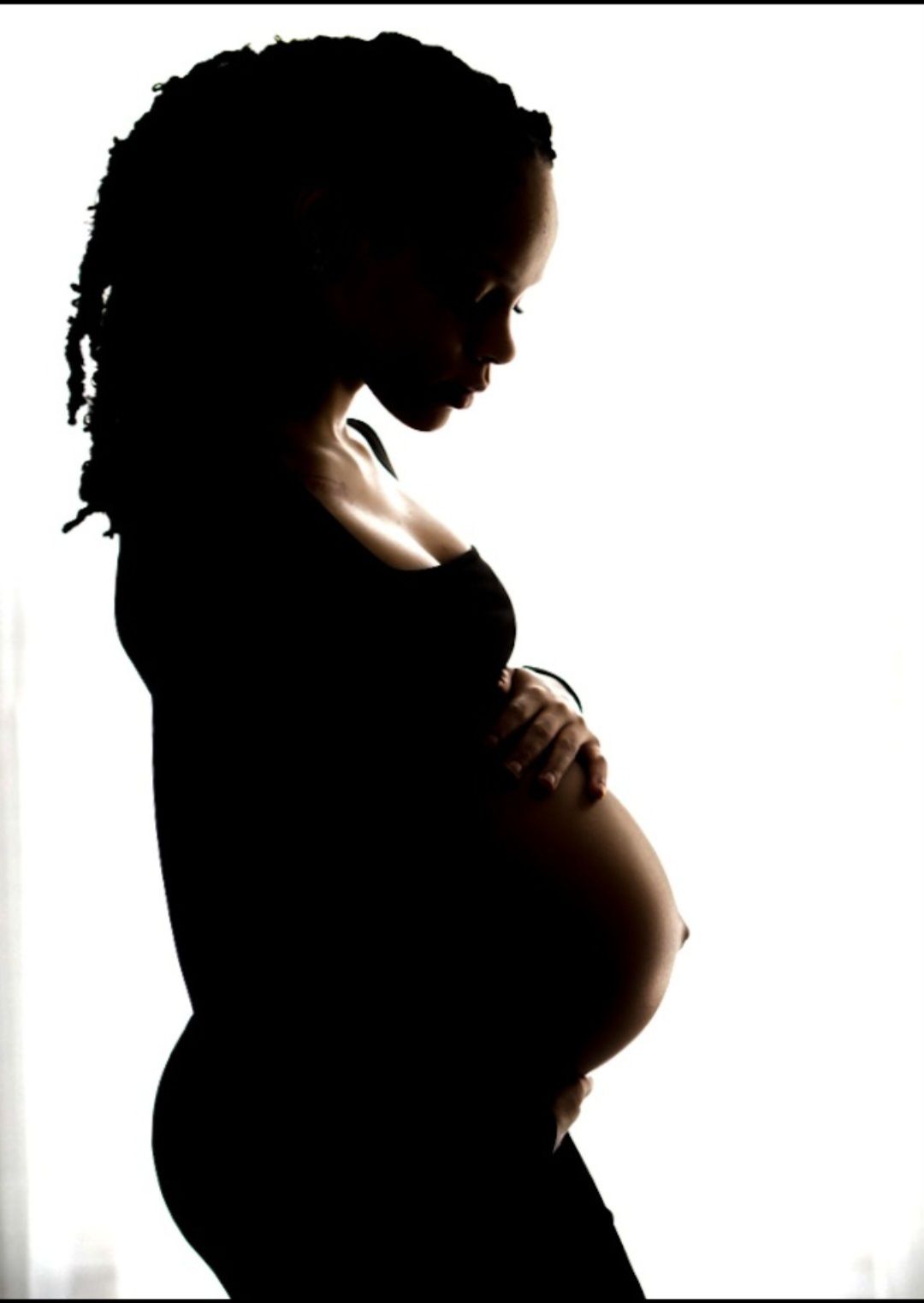Dramatic black silhouette of a pregnant woman in profile cradling her belly against a bright white background.