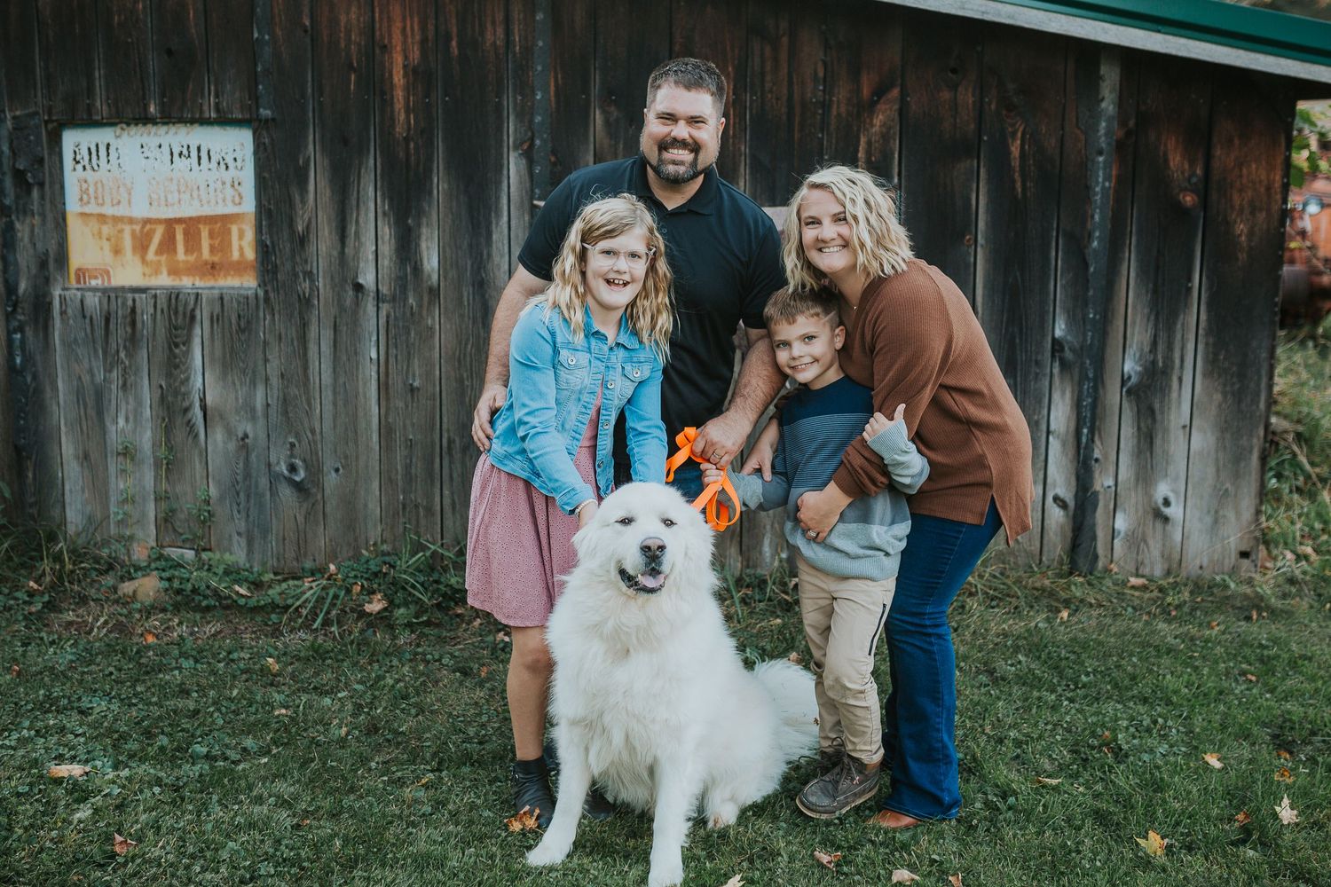 Happy family with white fluffy dog posing together in front of rustic barn.