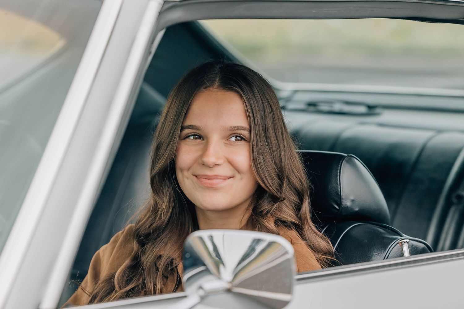 Smiling teenage girl with long brown hair sitting in a classic vintage car looking out the window.