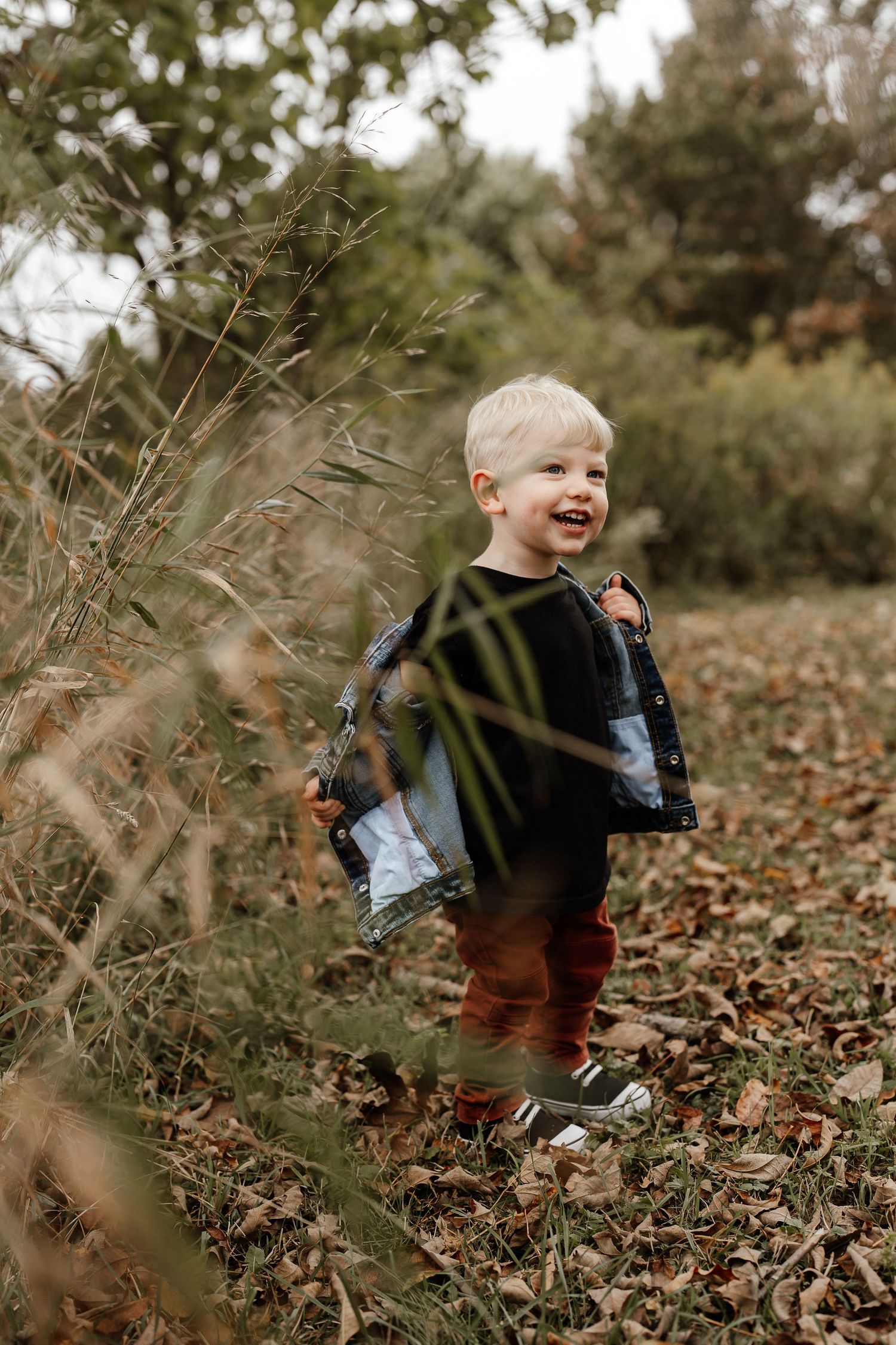 Laughing child plays outdoors in fallen autumn leaves surrounded by tall grass and trees.