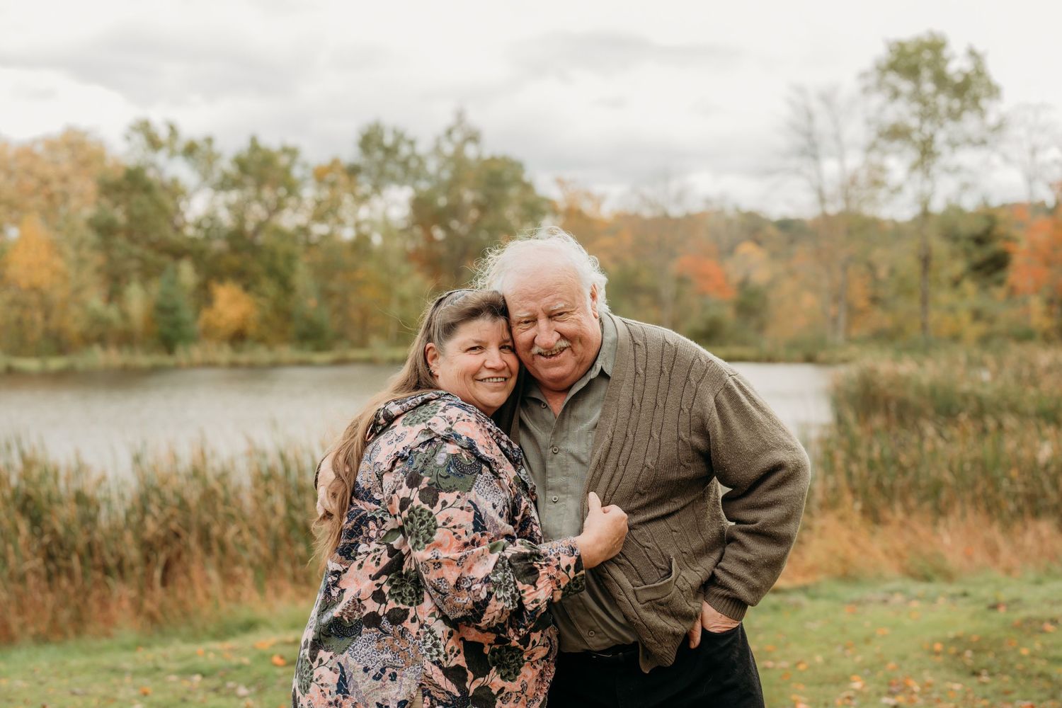 A couple embracing and laughing together near a pond with fall colors in the background.