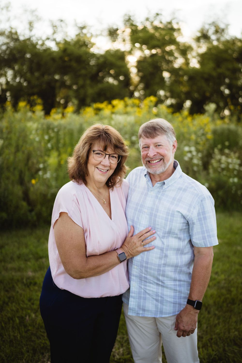 A happy couple poses together in a meadow at sunset wearing casual summer attire.