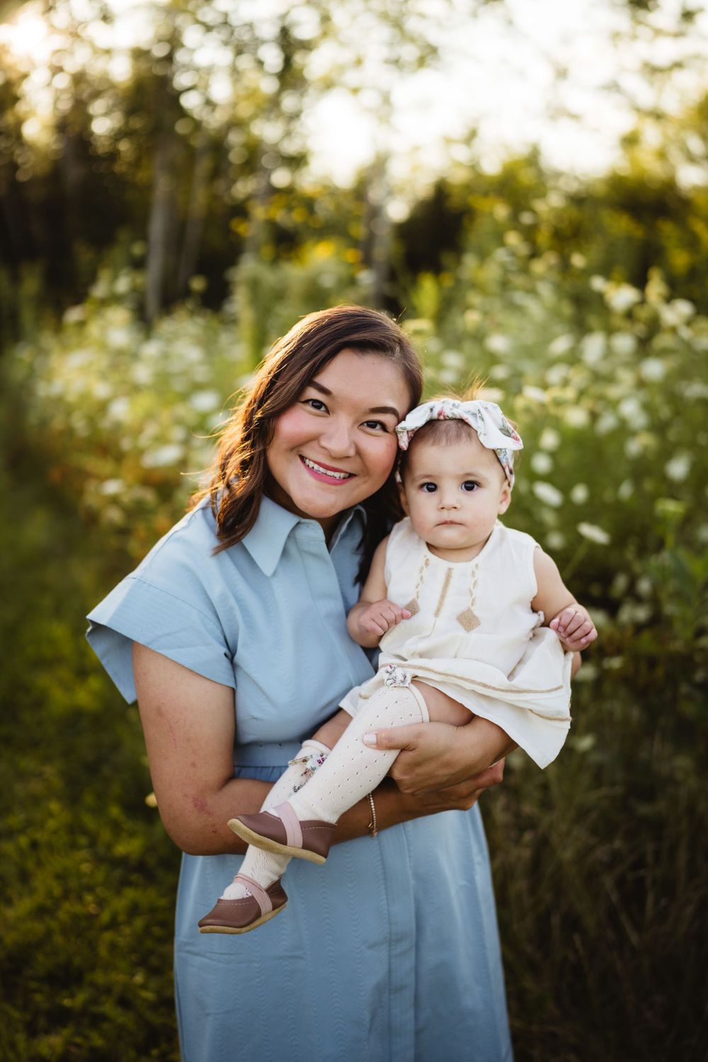 Baby in white dress and bow sits on lap of woman in blue dress during golden hour outdoor photoshoot.
