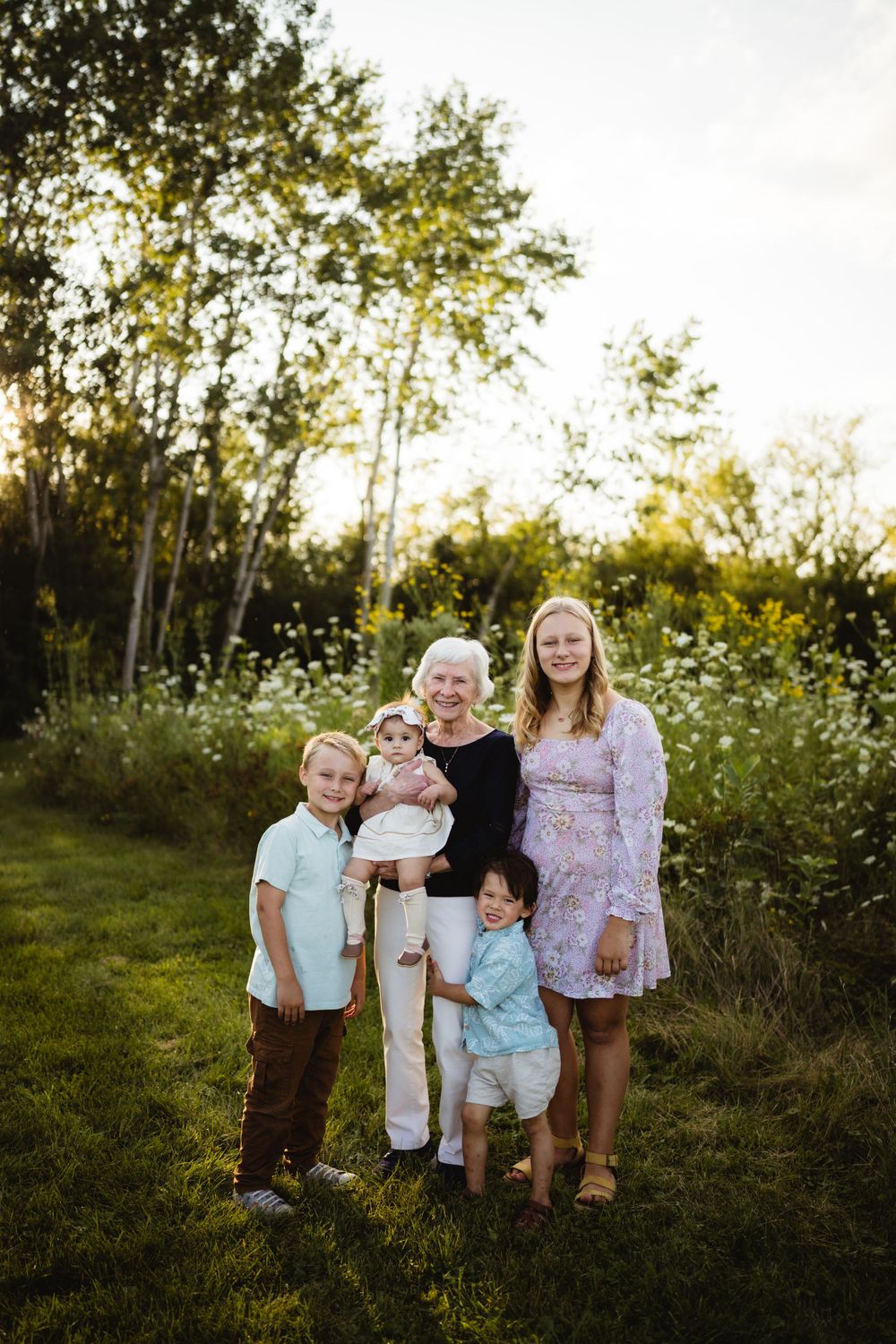 Family group poses together in casual outfits against natural wildflower backdrop at sunset.