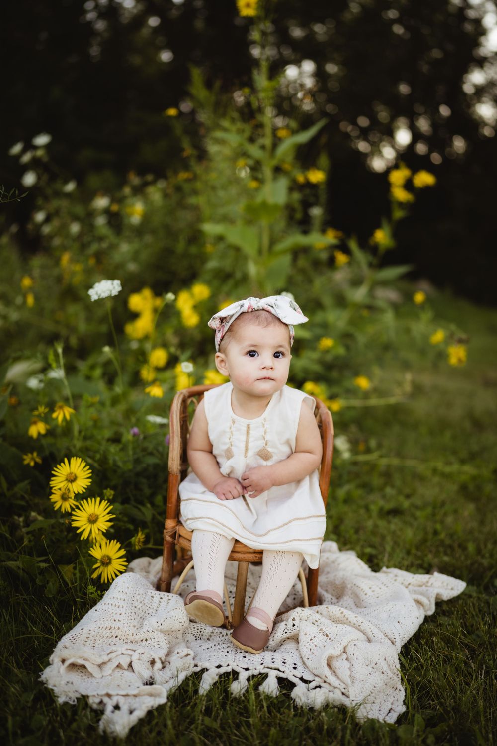 Baby in white dress and bow sits on lace blanket surrounded by yellow wildflowers.