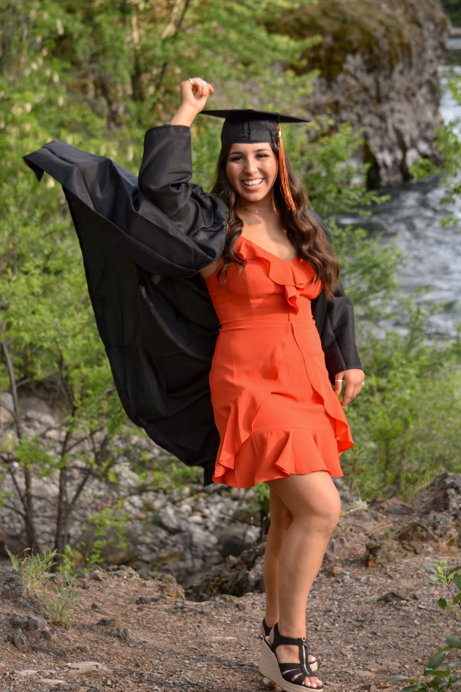 Happy young woman in graduation cap and gown over an orange dress celebrating outdoors near a river.