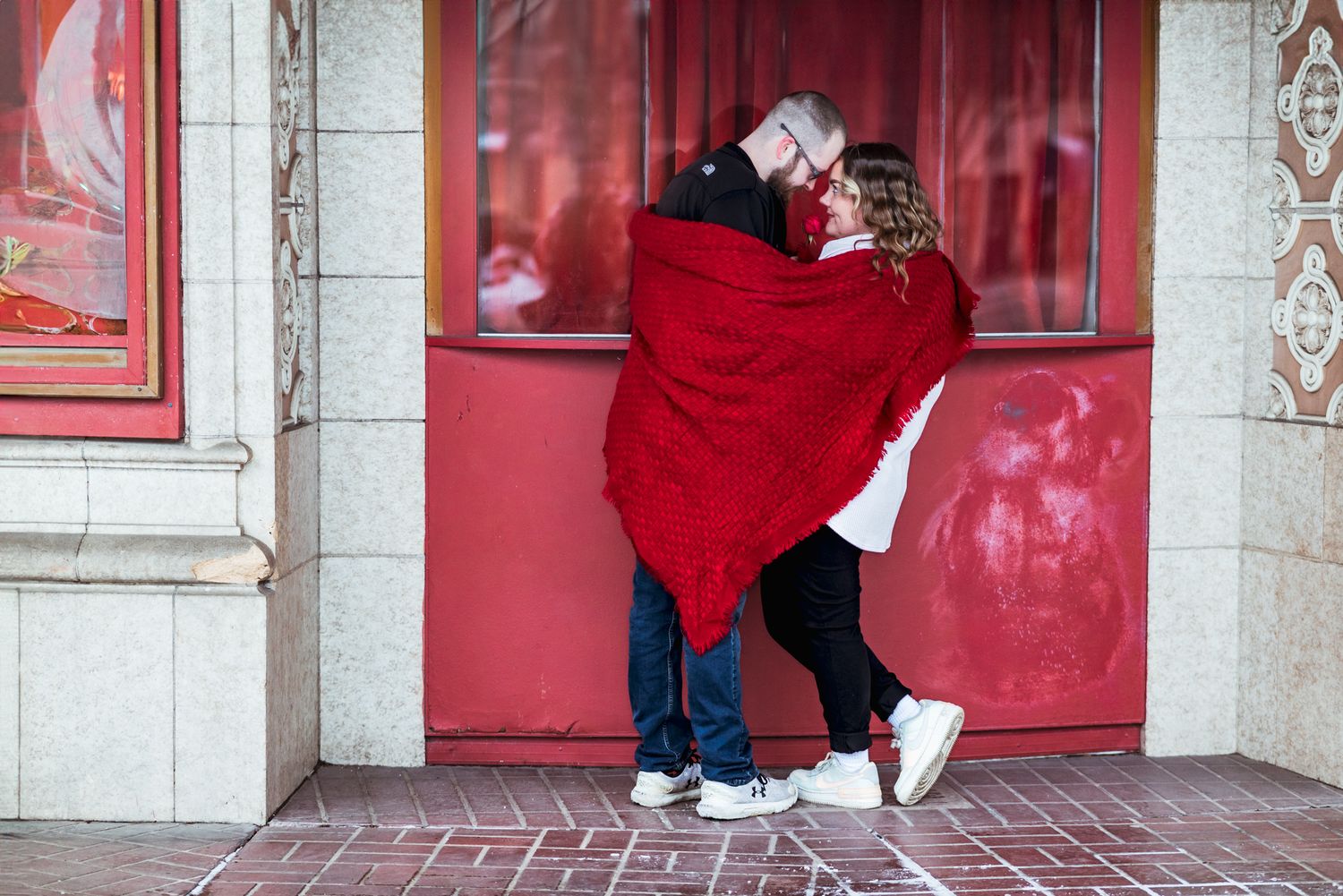 Two people wrapped in a cozy red blanket embrace while standing near a red door in an urban setting.