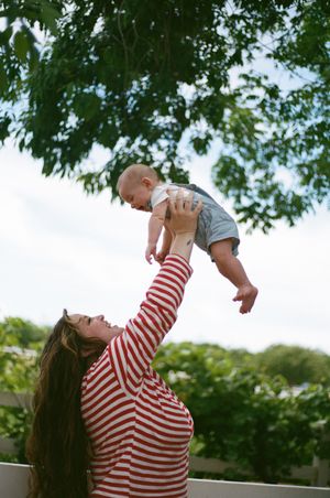 Woman in striped shirt lifts baby in air against leafy green background.