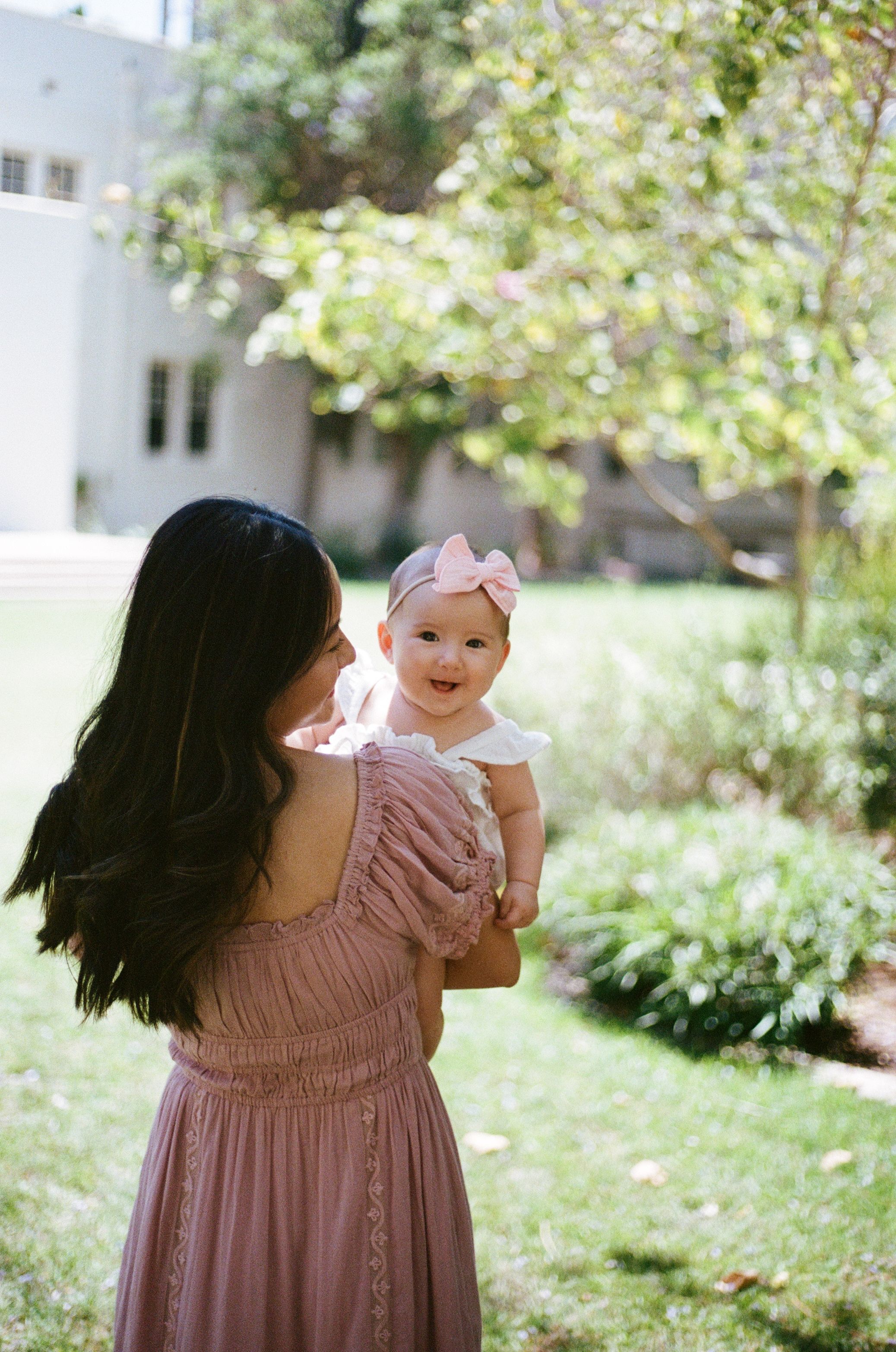 A mother in a pink dress holds her smiling baby in a sunlit garden setting.