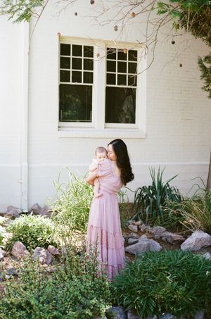 A mother and baby stand together in front of a white building with garden plants.