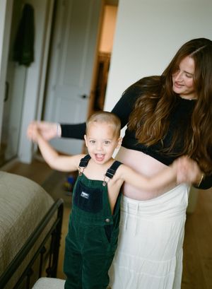 Candid moment of parent and young child in green overalls dancing together in a sunlit living room.
