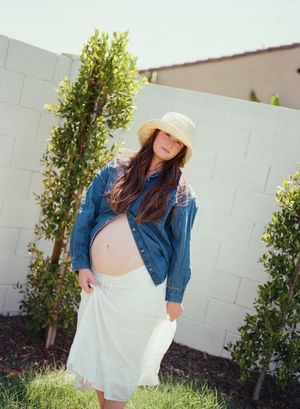 Pregnant person in a denim jacket and sun hat poses outdoors against a white wall with green shrubs.