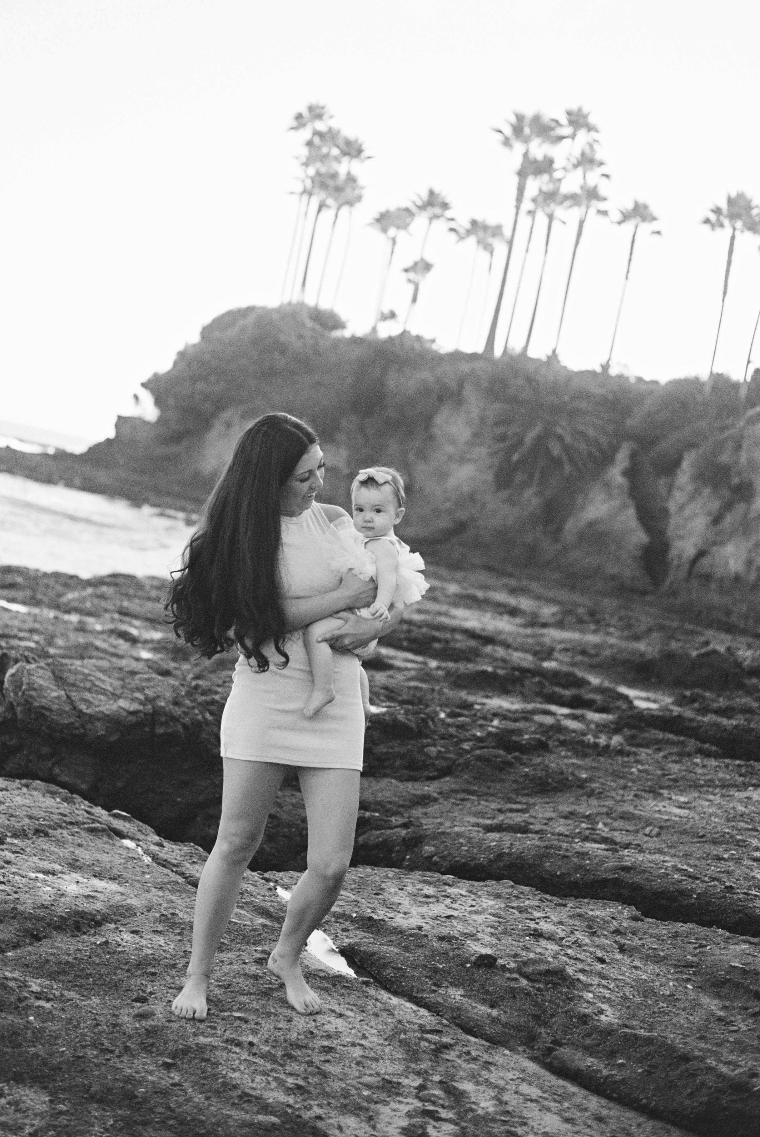 Parent and baby pose on rocky beach with palm trees in background.