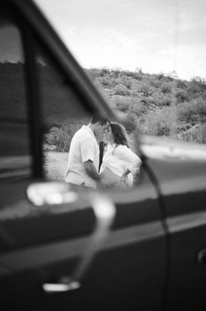 A romantic black and white photo taken through a car window shows a couple sharing an intimate moment in a desert landscape.