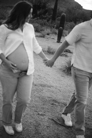 A pregnant woman in white holds hands with a companion while walking along a desert trail with saguaro cacti.