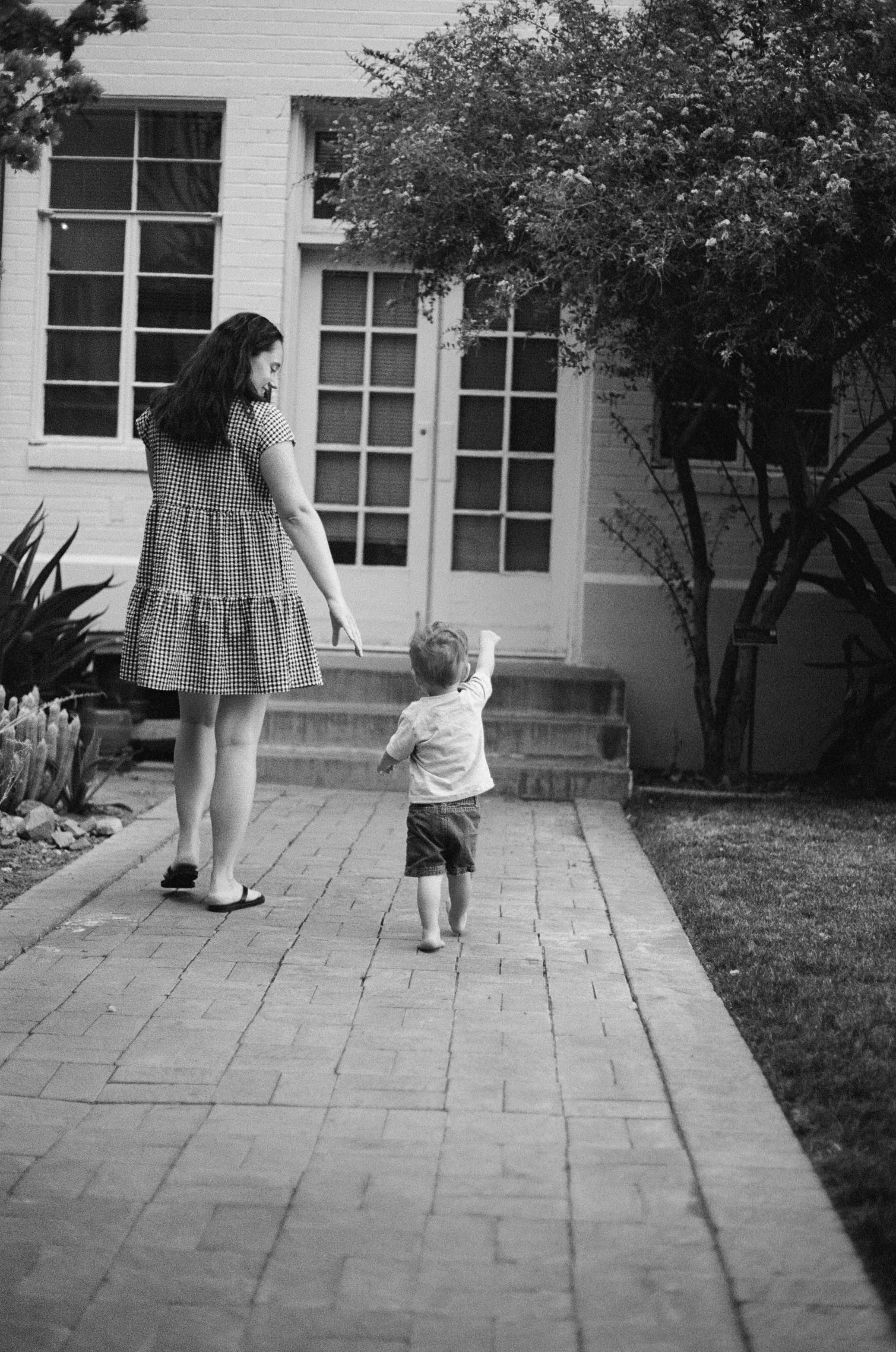 A black and white image shows a touching moment between parent and child walking along a garden path.