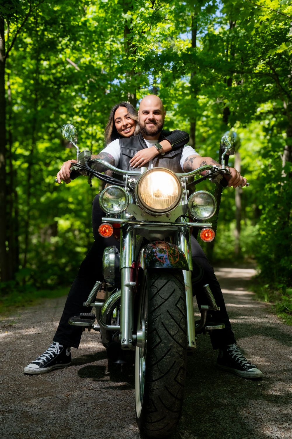 Couple poses on vintage motorcycle in lush green forest setting.