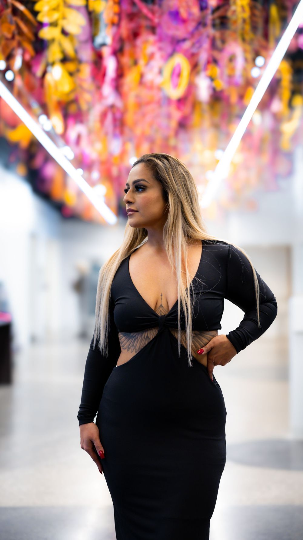 Figure in black dress poses against colorful floral installation with neon lighting.