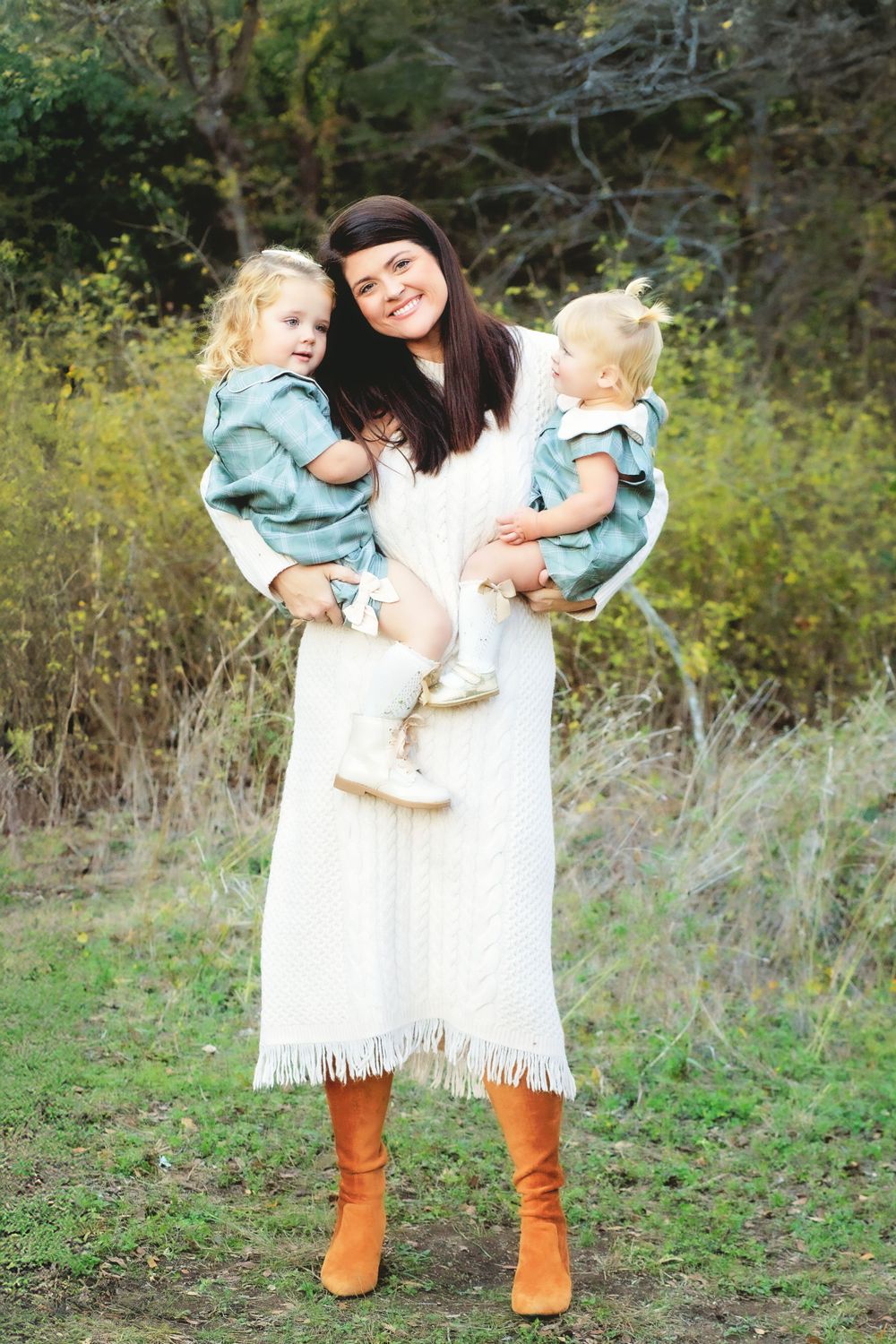 Standing outdoors in a white fringe dress and tan boots while holding two young children against a natural background.