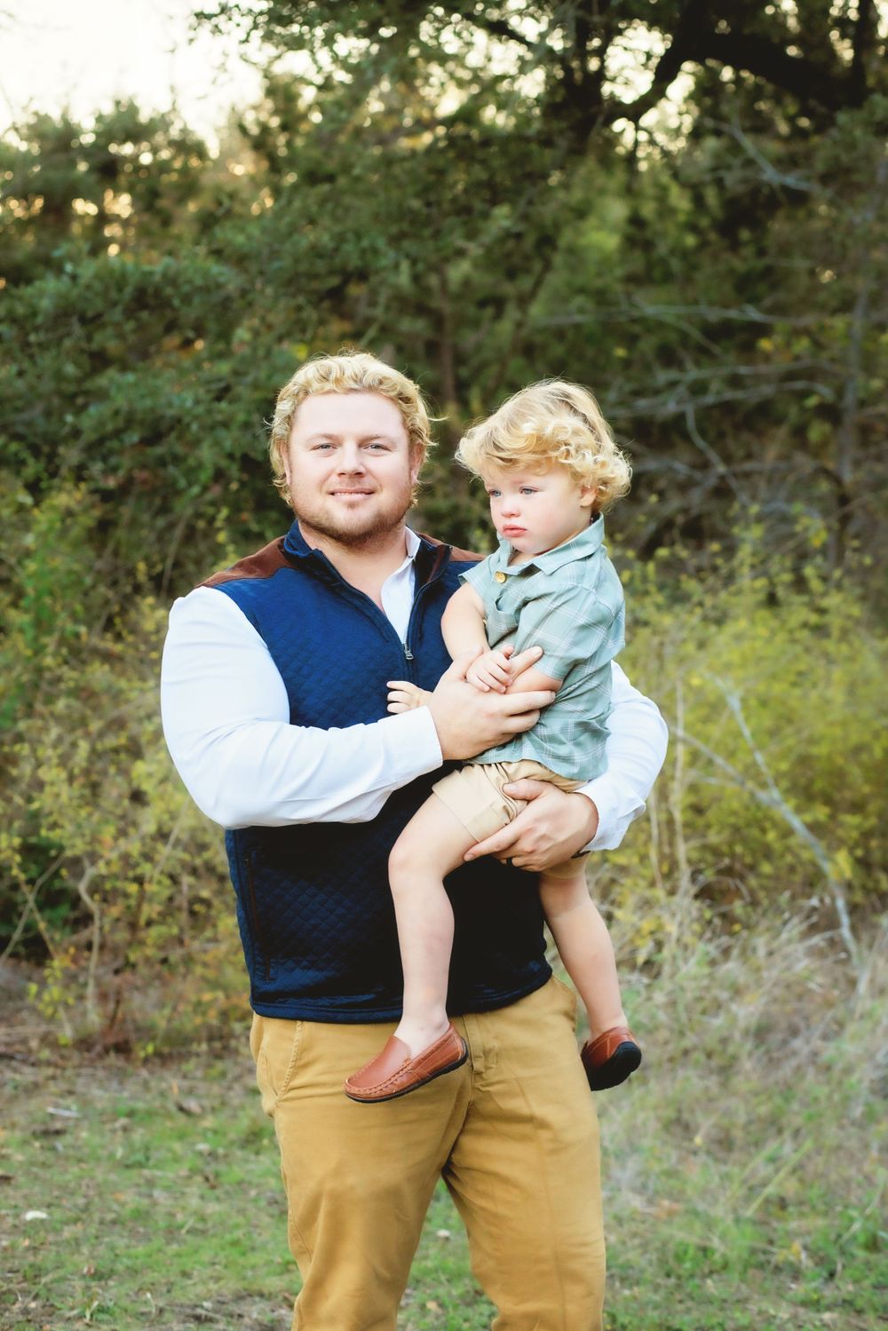 A figure in a navy vest and khaki pants holds a young child wearing light clothing outdoors in a natural setting.