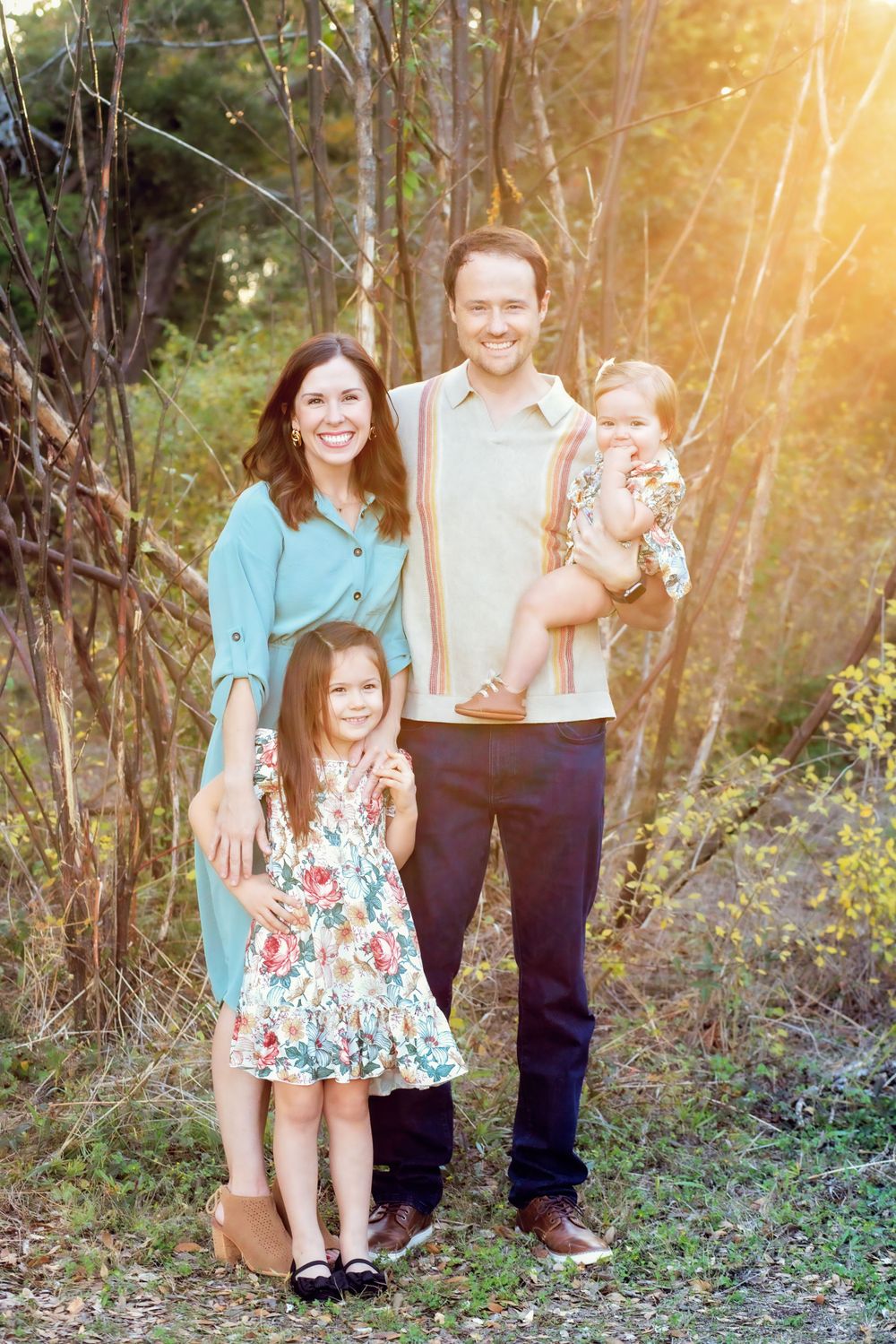 A family poses together outdoors in a wooded setting during golden hour with sunlight filtering through trees.