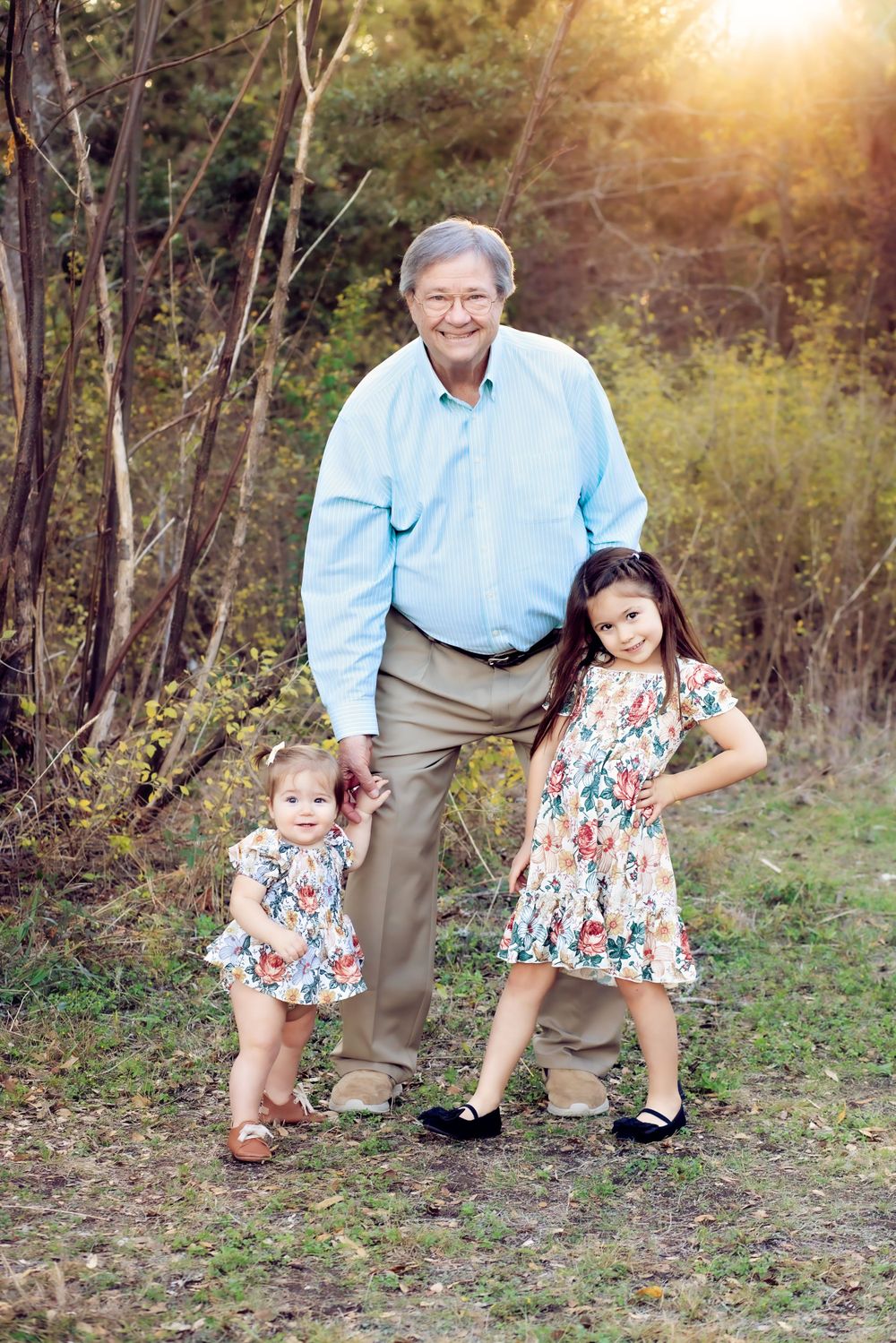 Two young girls in floral dresses pose outdoors with an elderly person in a light blue shirt at sunset.