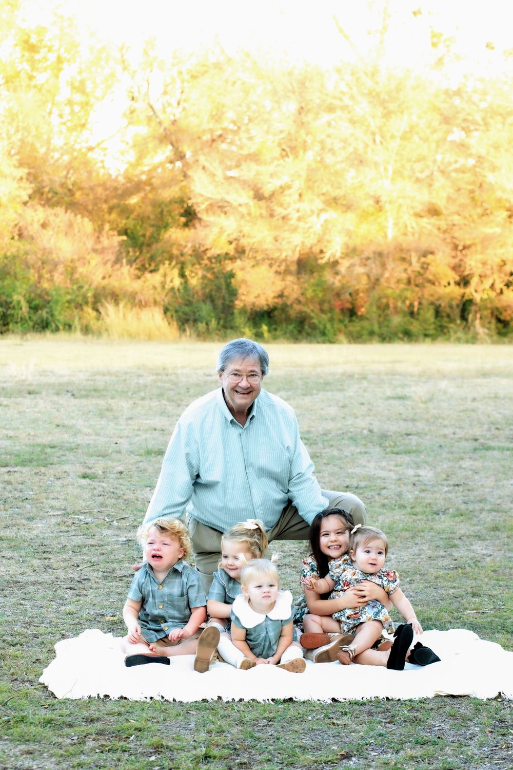 A group sits together on a white blanket in a grassy field with autumn trees in the background during golden hour.