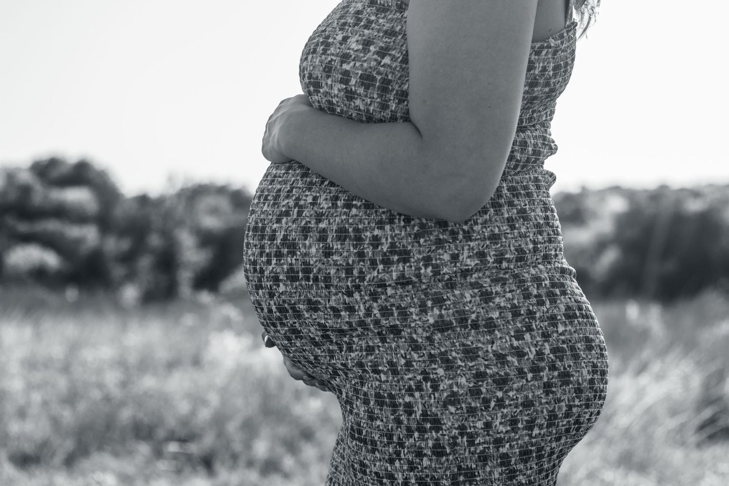 Black and white side profile of a pregnant belly in a floral dress standing outdoors in nature.