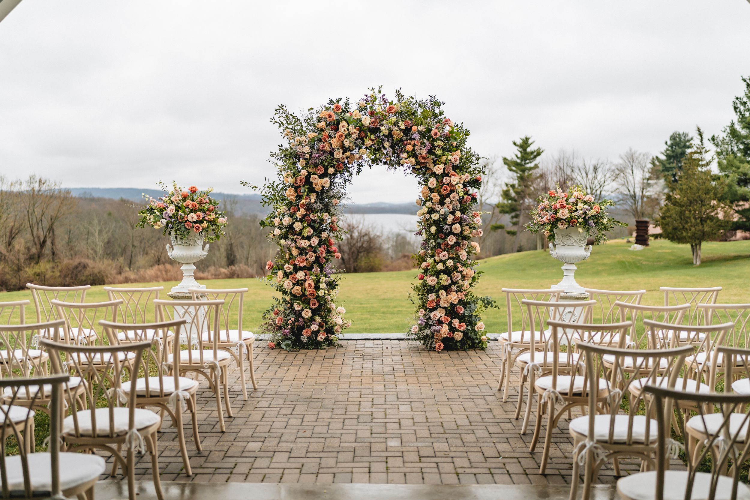 Beautiful outdoor wedding ceremony setup with floral arch and white chairs on stone patio overlooking scenic landscape.