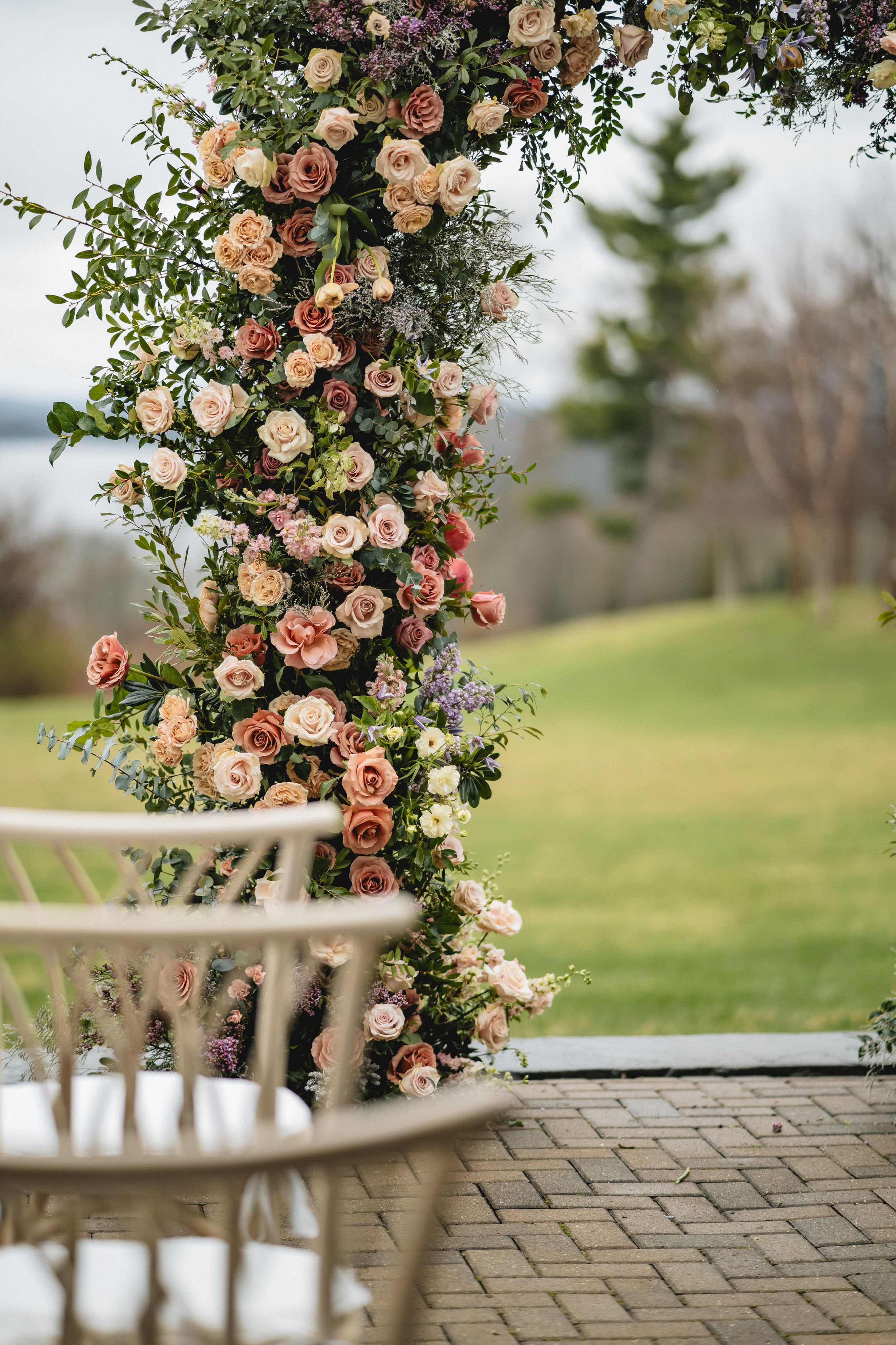 A floral wedding arch with pink and cream roses cascades over outdoor seating with a scenic landscape view.