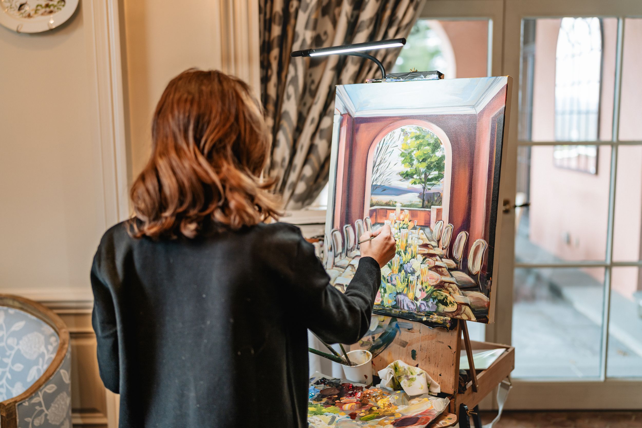 An artist works at their easel painting artwork in a well-lit room with natural light streaming through French doors.