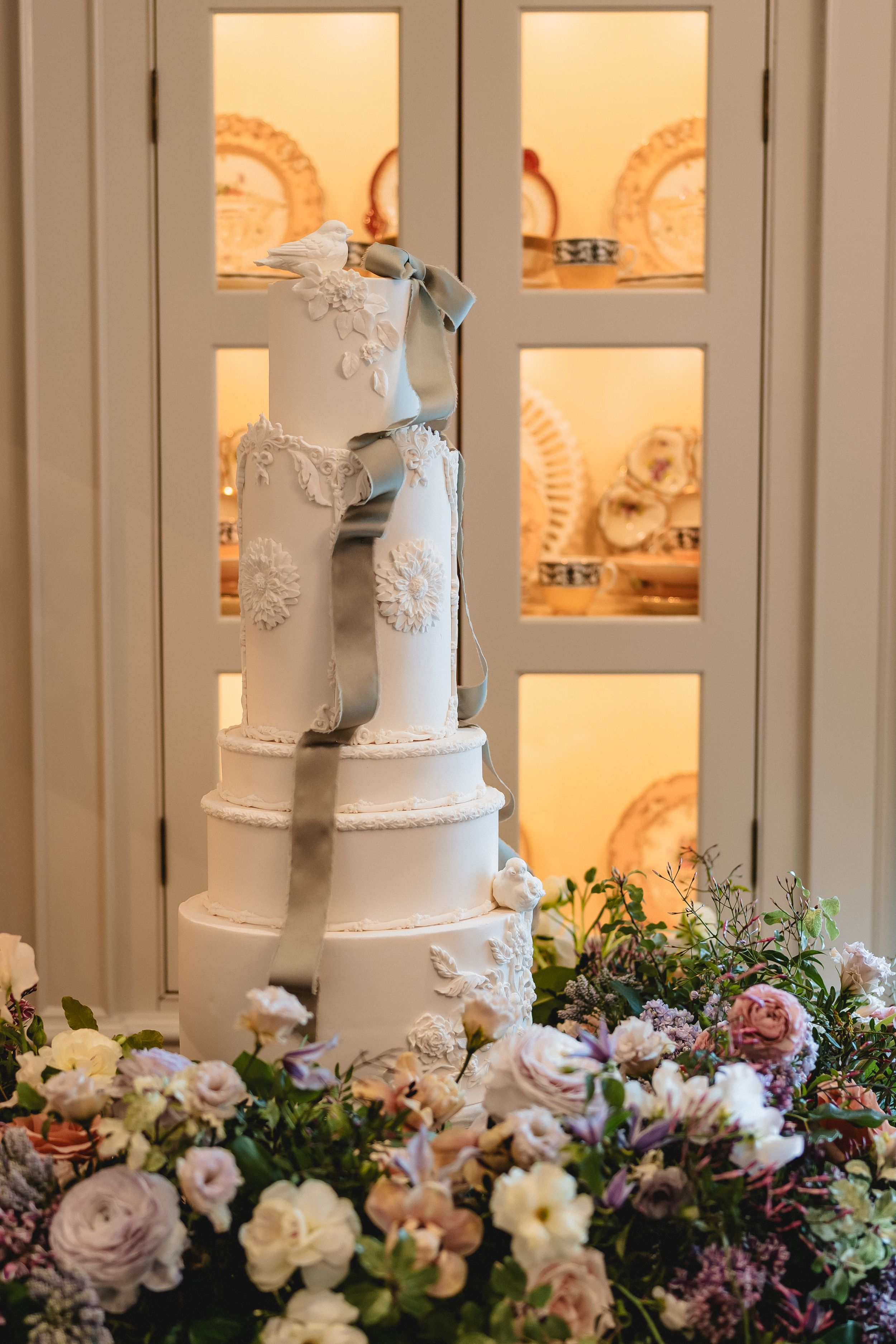Elegant white tiered wedding cake decorated with delicate lace details and surrounded by floral arrangements.