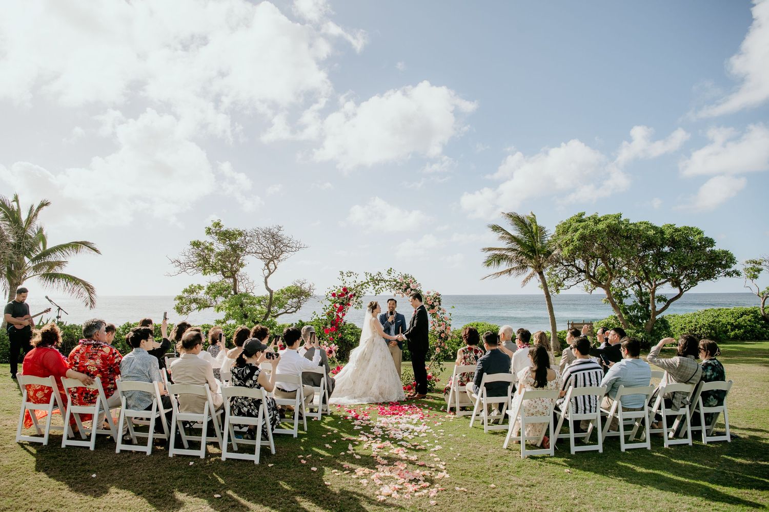 Beautiful beachfront wedding ceremony with guests seated on white chairs under palm trees and blue tropical skies.