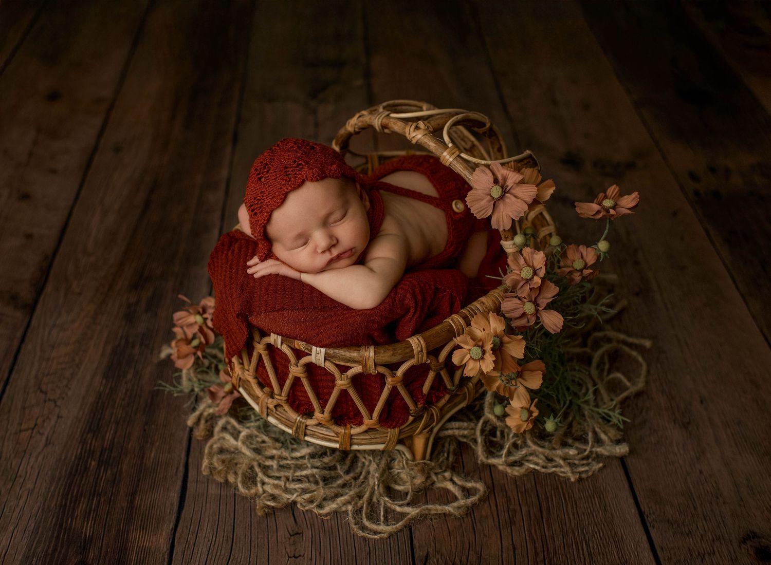 A sleeping newborn nestled in a red woven basket surrounded by delicate dried flowers on a wooden floor.