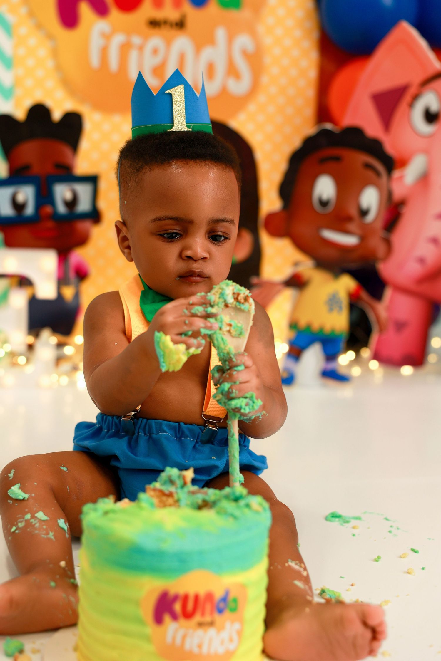 A one year old celebrates their first birthday with a green and yellow cake at a Kurb Friends themed party celebration.