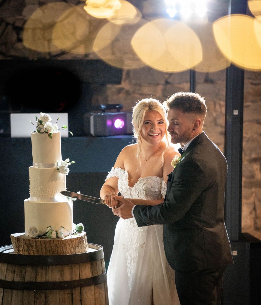Elegant white wedding cake displayed on rustic barrel with couple dancing in background.