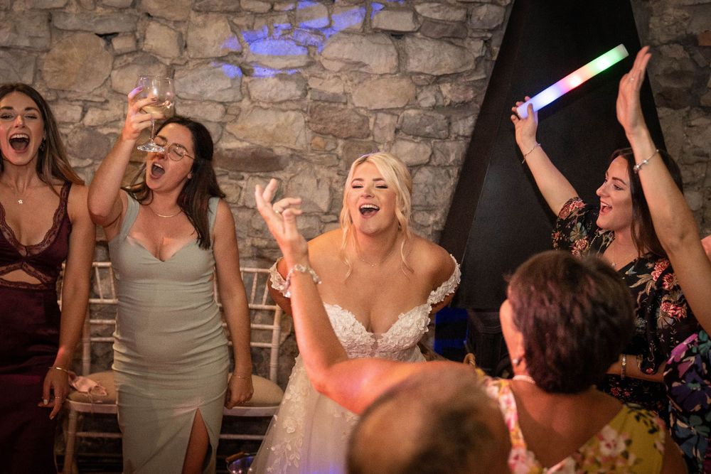 Wedding reception party with guests dancing beneath stone walls and colored lighting.