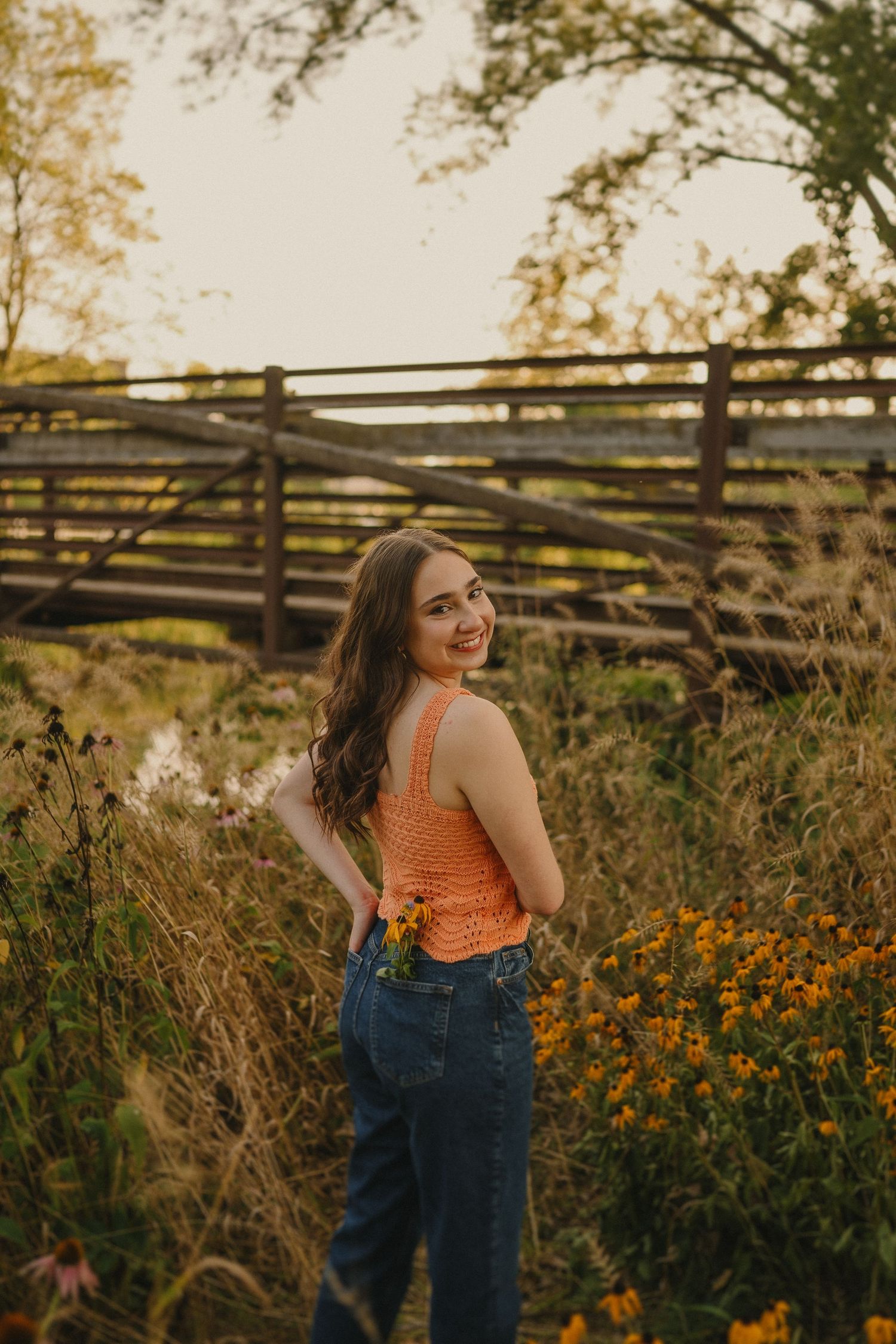 A golden hour portrait in an orange top and jeans poses by a rustic wooden fence.