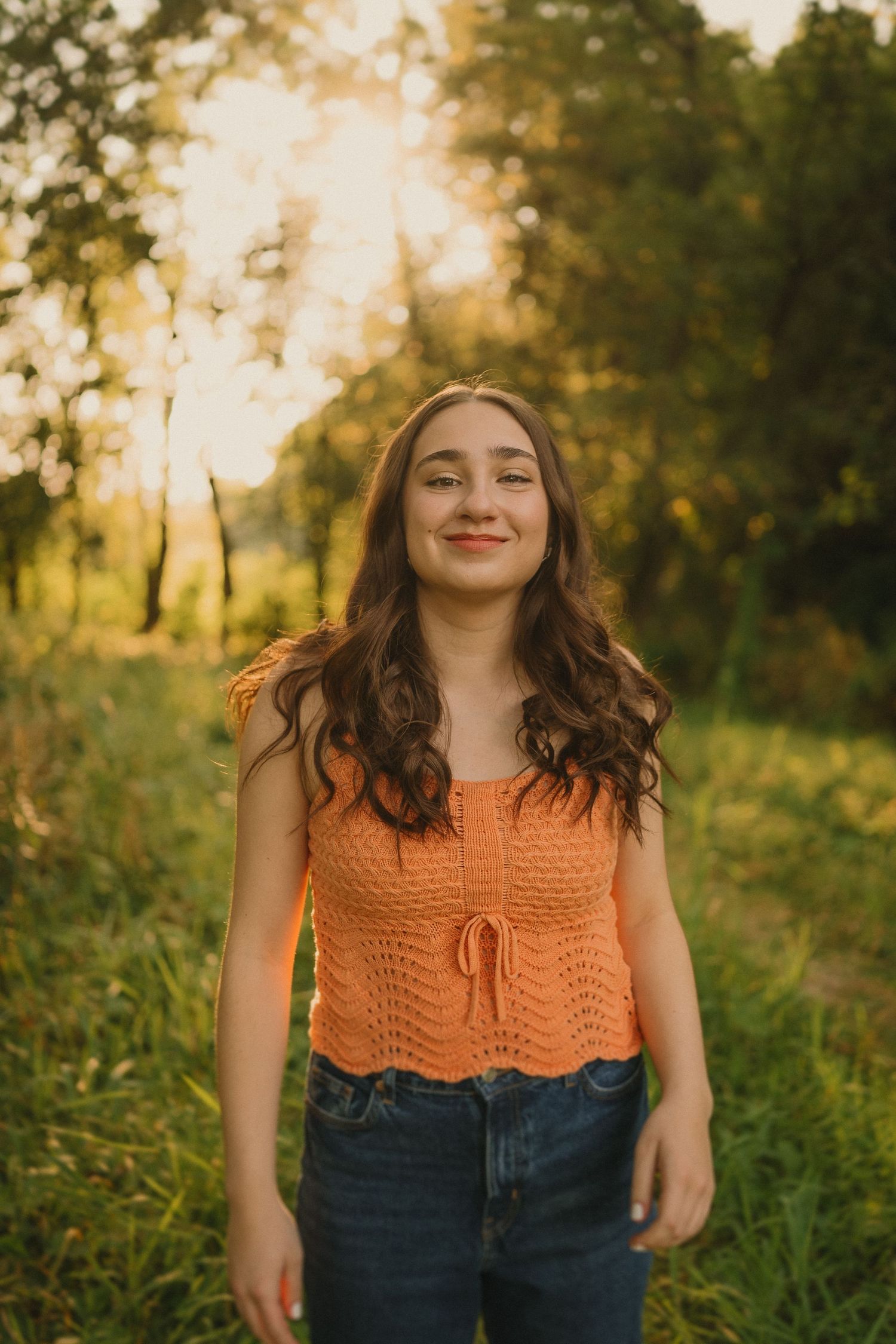 A candid outdoor portrait at golden hour showing someone in an orange knit top and jeans surrounded by lush greenery.